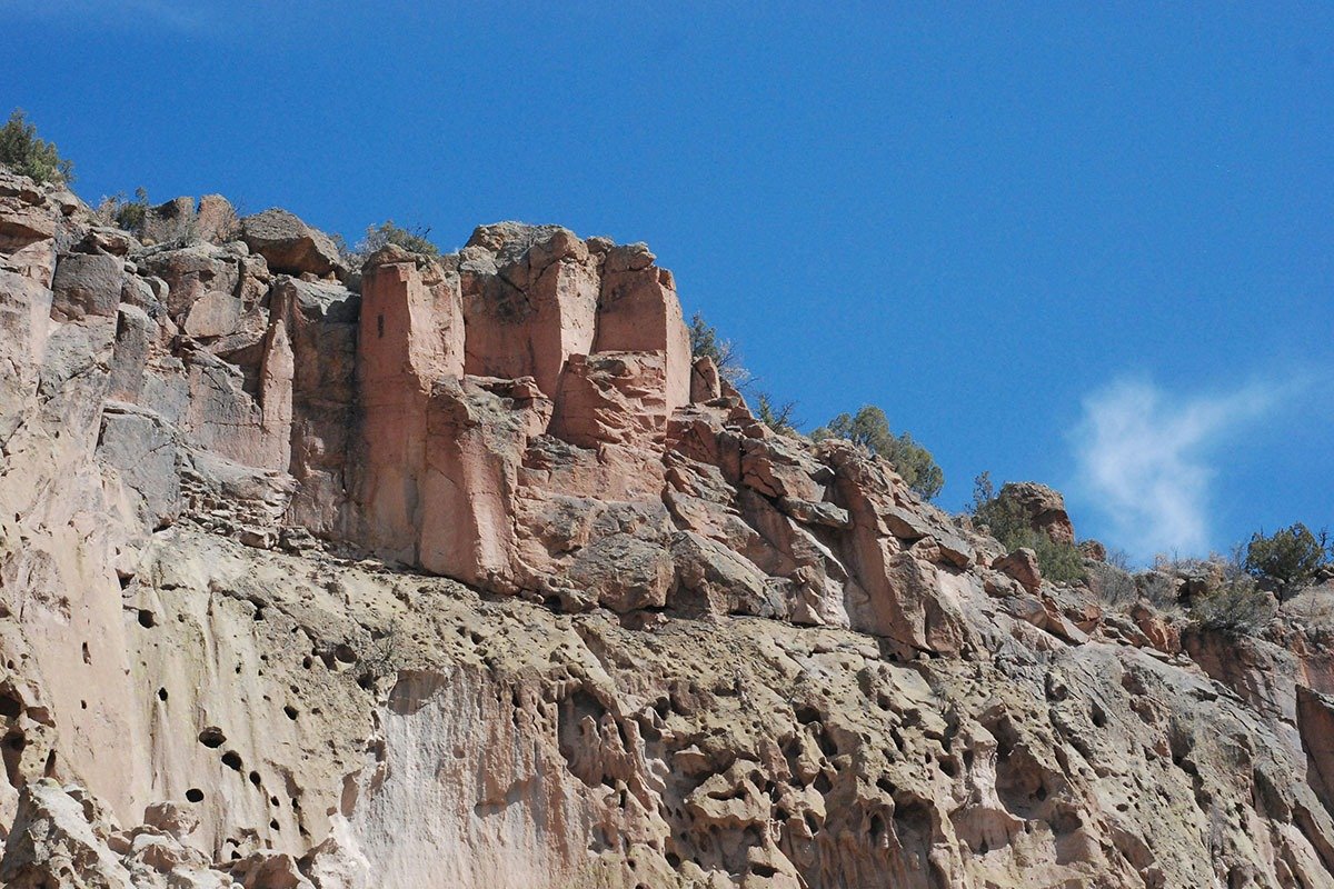 A rugged cliff featuring layered rocks in shades of pink and gray under a clear blue sky, with sparse vegetation at the top.