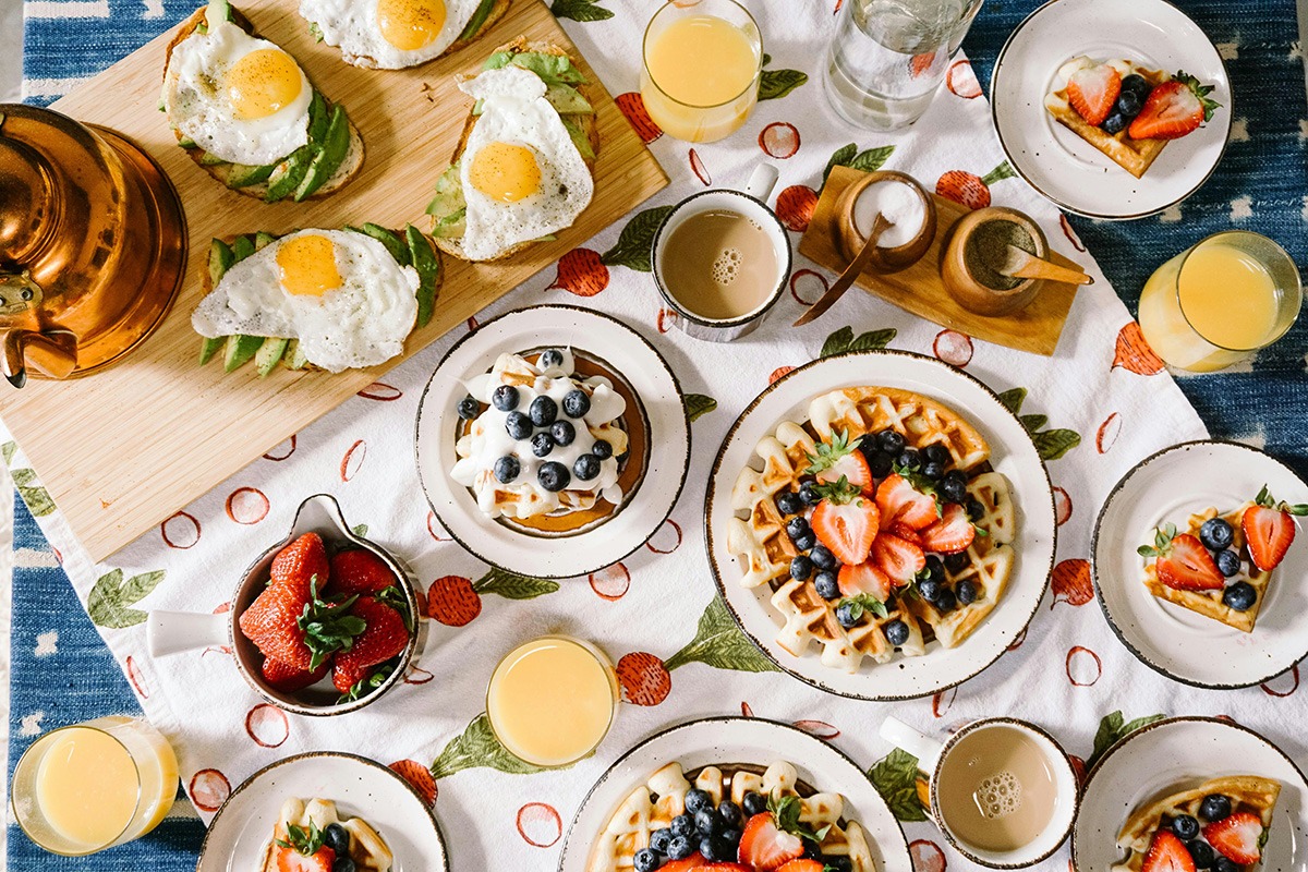 A vibrant breakfast table setting featuring plates of waffles topped with strawberries and blueberries, pancakes with cream and blueberries, avocado toast with fried eggs, and fresh strawberries. Beverages include orange juice and coffee, all arranged on a patterned tablecloth.