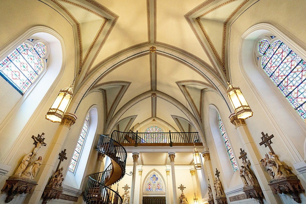 Interior view of a church with a high vaulted ceiling, featuring stained glass windows, ornate light fixtures, and a decorative spiral staircase leading to a balcony, alongside statues and wooden crosses adorning the walls.