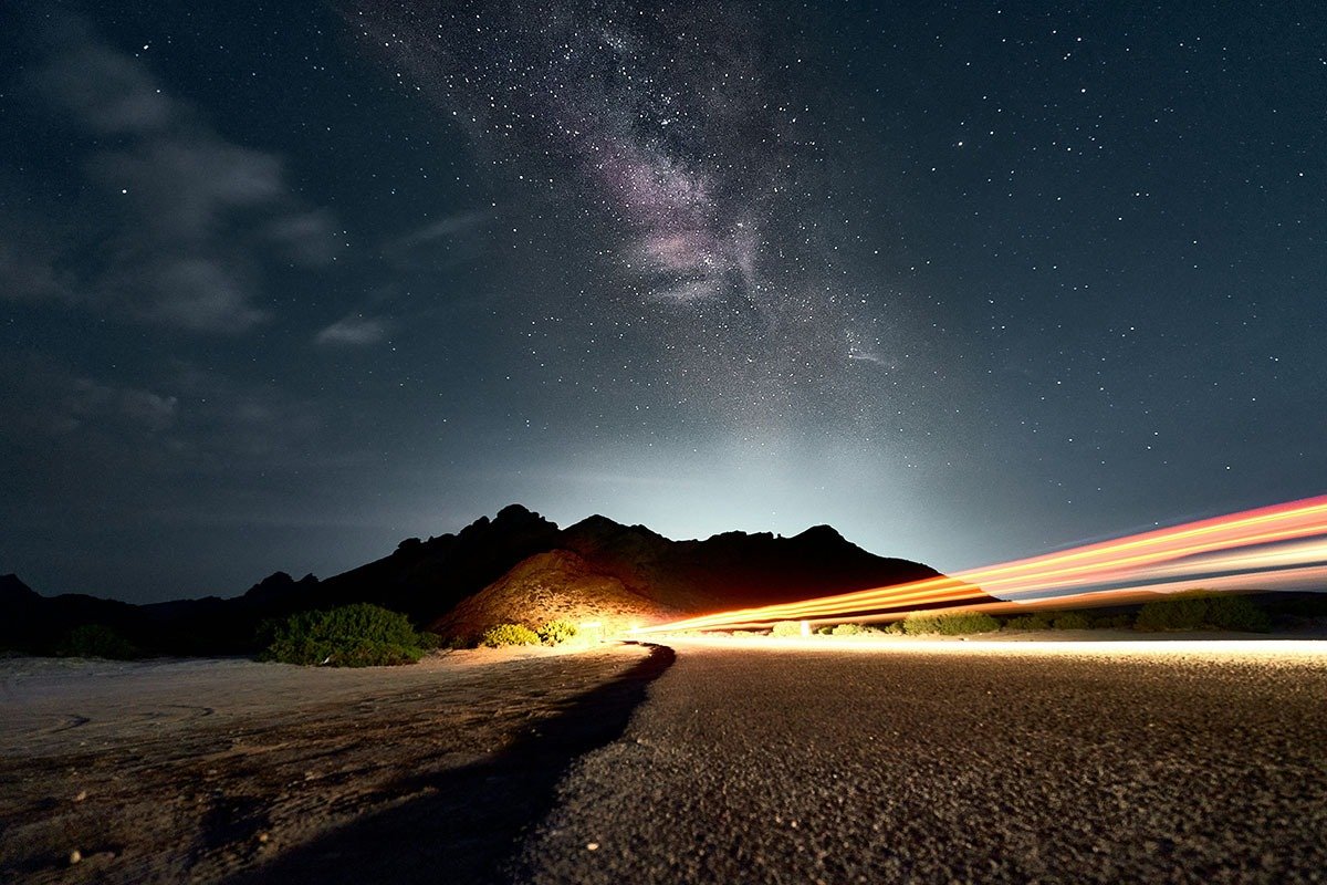 A winding road under a starry night sky, with a visible Milky Way stretching across the horizon, and the silhouette of mountains in the background. Bright streaks of light from a passing vehicle illuminate the scene.
