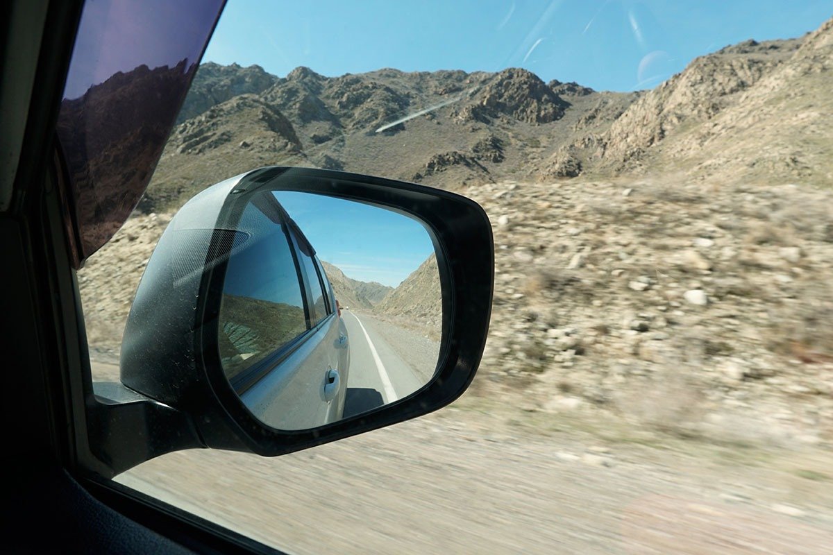 A close-up view of a car's side mirror reflecting a mountainous landscape and a winding road, with a clear blue sky above.