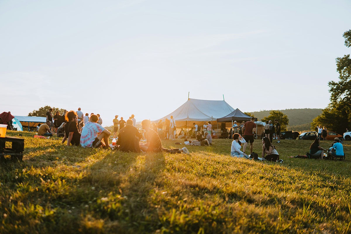 A vibrant outdoor gathering at sunset, featuring a diverse crowd of people sitting and socializing on a grassy field, with a large white tent in the background and hills in the distance.
