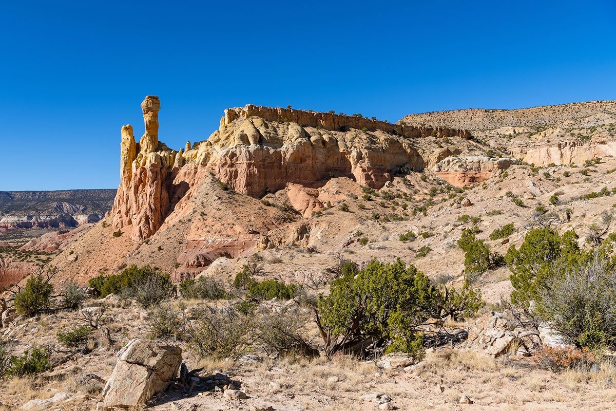A rocky landscape features a tall, narrow rock formation with a light-colored top, surrounded by multi-hued cliffs and sparse vegetation under a clear blue sky.