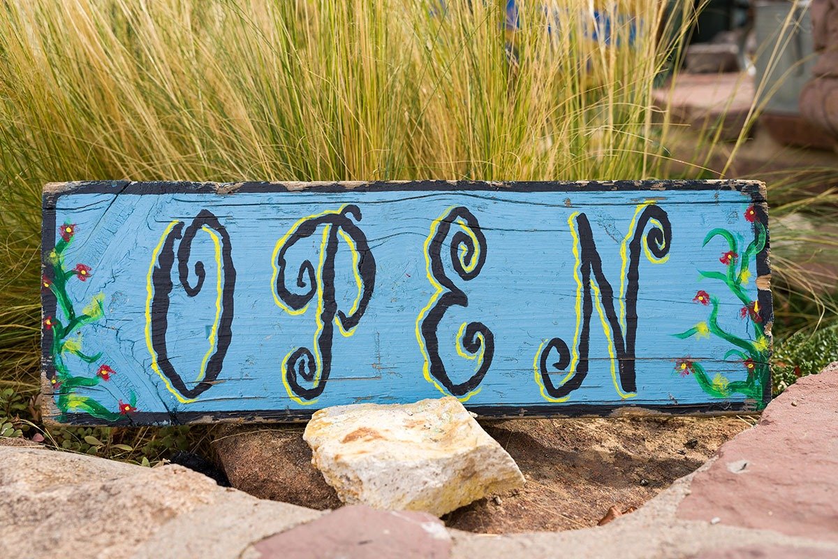 A blue wooden sign painted with the word 'OPEN' in black, decorative letters, surrounded by small floral designs, placed on natural stones with tall grass in the background.