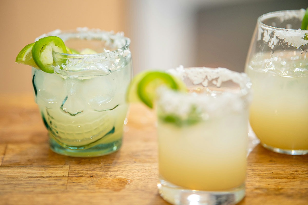 Three cocktails are displayed on a wooden surface: one served in a skull-shaped glass with ice and garnished with lime and jalape&ntilde;o slices, and two others in round glasses with salted rims, one containing a light-colored drink and the other a darker beverage.