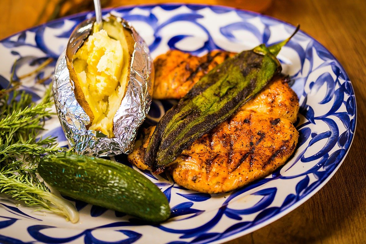 A plated meal featuring grilled chicken breast, a baked potato wrapped in aluminum foil filled with butter and seasoning, a grilled green pepper, and fresh herbs, all arranged on a decorative blue and white patterned plate.