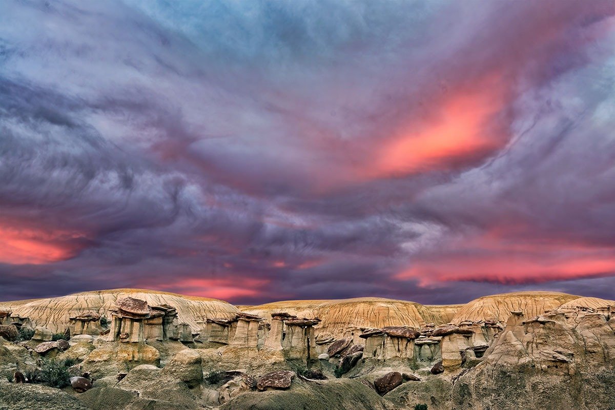 A dramatic landscape featuring eroded rock formations in varying shades of tan and gray, under a colorful sky with vibrant pink and purple clouds at sunset.