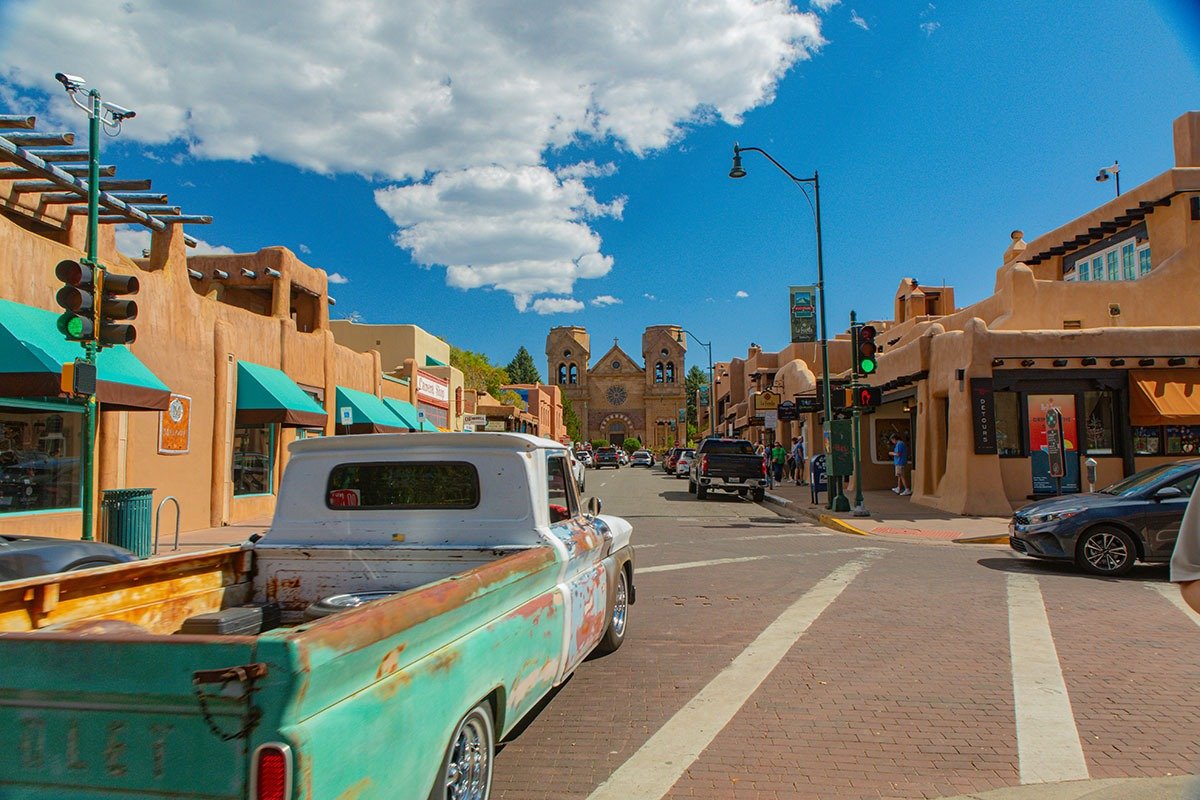 A vintage turquoise and white truck drives down a bustling street lined with adobe-style buildings featuring green awnings and shops. In the background, a historic church with twin towers is visible under a bright blue sky with puffy clouds. Traffic signals and various vehicles populate the scene, capturing a lively atmosphere.