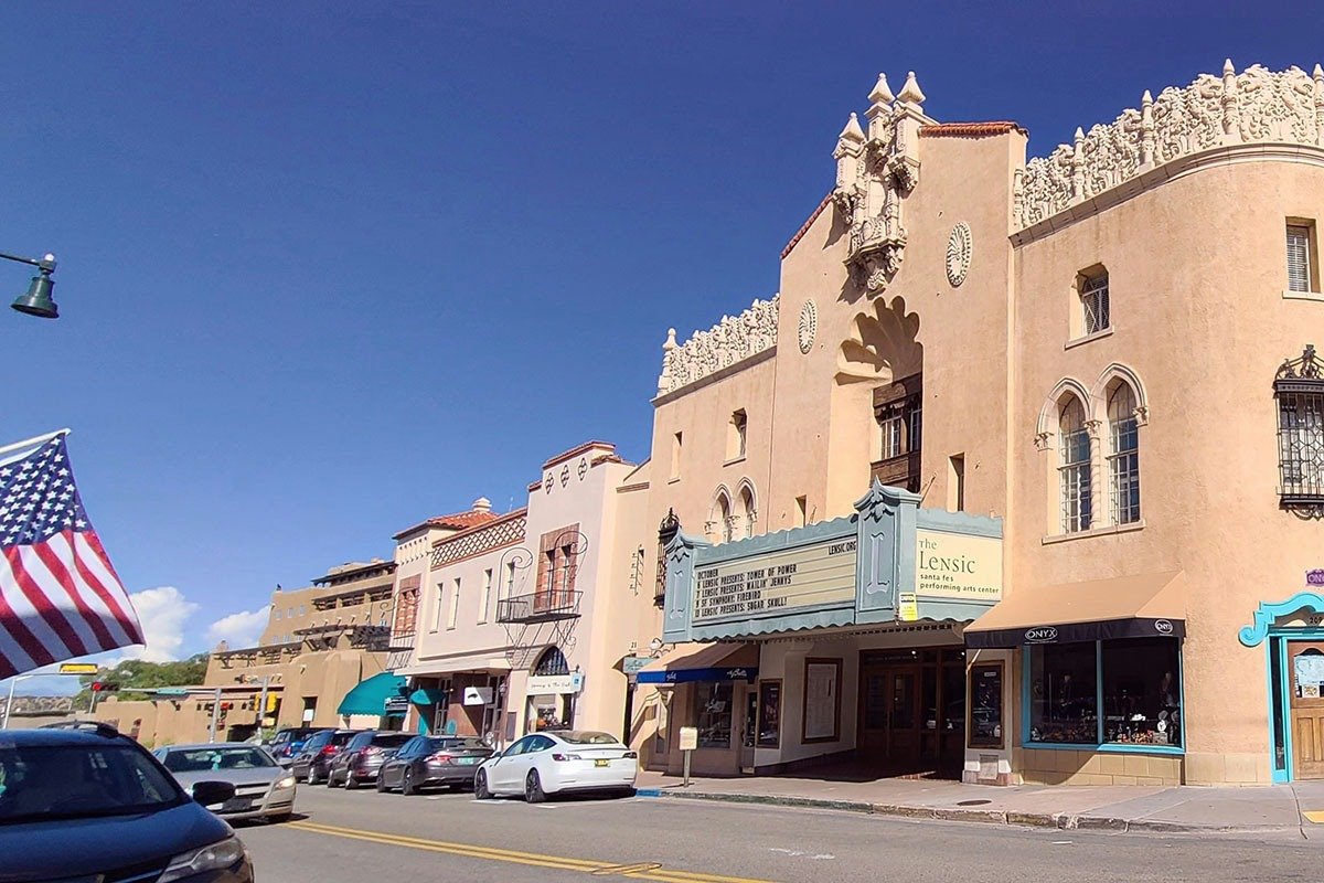 A street view of the Lensic Performing Arts Center, featuring its decorative facade and marquee, surrounded by southwestern-style buildings and parked cars, with an American flag in the foreground under a clear blue sky.