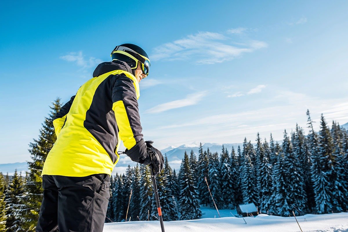 A skier in a bright yellow and black jacket stands on a snowy slope, holding ski poles and looking out at a panoramic view of snow-covered mountains and evergreen trees under a clear blue sky.