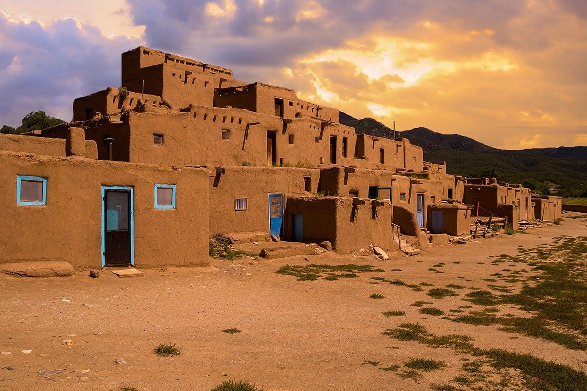 A landscape image of multi-storied adobe buildings with turquoise doors under a dramatic sunset sky, surrounded by open ground and distant mountains.