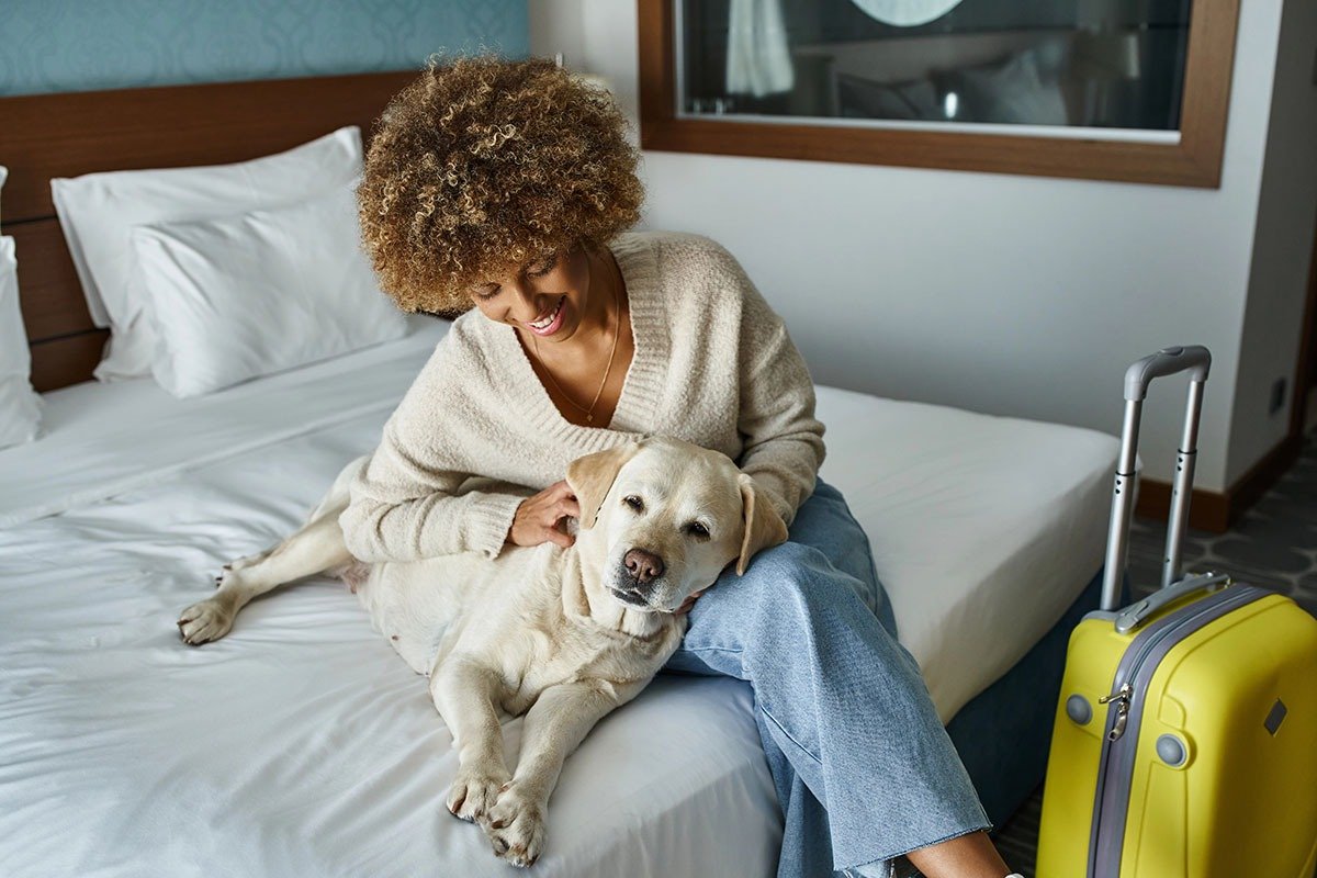 A woman with curly hair sits on a bed, smiling as she pets a Labrador dog. A yellow suitcase is beside them, and the room features soft bedding and a window in the background.