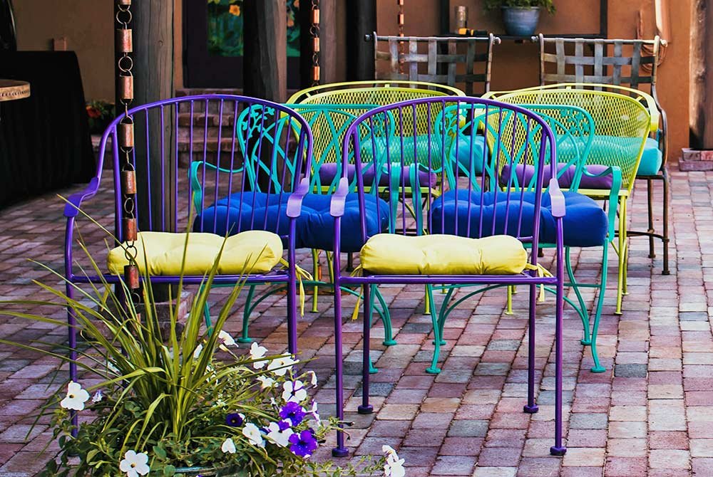 A collection of brightly colored metal chairs in purple, blue, and yellow, arranged around a paved outdoor area, with surrounding greenery and flowers.